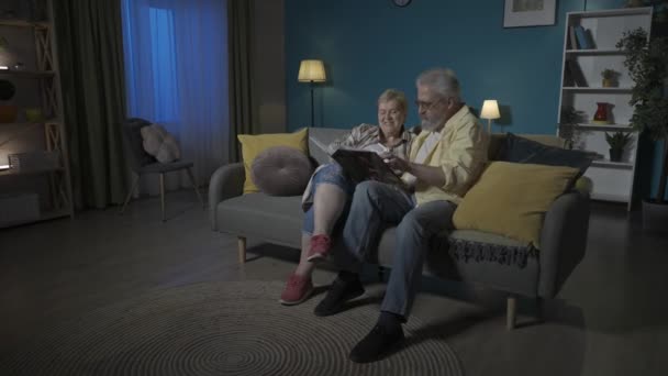 在画面中 一对老夫妻坐在公寓的沙发上 靠着一面蓝色的墙 他们正在看 翻阅一本相册 Hdr Bt2020 Hlg Material — 图库视频影像