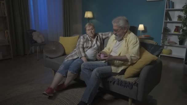 写真では お年寄りのカップルが部屋のソファーに座っています 男性は枕の下に隠されたピンクの箱に手を伸ばします 女性のための贈り物 彼女を驚かせたい 彼女は見ていないか知っている Hdr Bt2020 Hlg — ストック動画