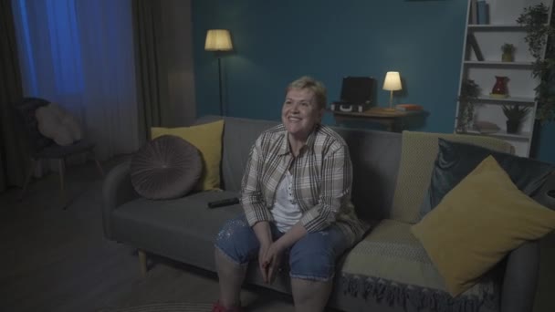 写真では 高齢の女性がアパートのソファーに座っています まるで映画や番組を見ているかのようだ 彼女は幸せで 彼女は笑っている 彼女は面白いです Hdr Bt2020 Hlg材料について — ストック動画
