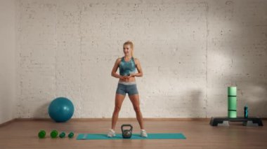 Evde kişisel spor eğitimleri. Spor giyimli bir kadın spor yapıyor. Sağlık ve sağlık reklamları konsepti. Atletik kadın fitness koçu çevrimiçi dersler için kettlebell ile çömelme hareketi yapıyor..