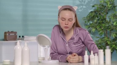 İpek pijamalı ve bantlı bir kadın tuvalet masasında otururken aynada kendine bakıyor. Bir kadın bir krem kavanozunu alır ve sorunlu cilt bölgelerine uygular.