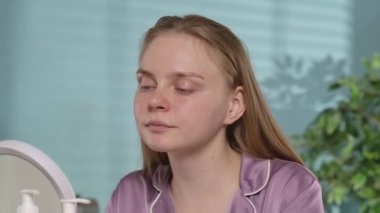 Derisi sorunlu genç bir kadın bir krem kavanozunu açar ve yüzüne sürer. İpek pijamalı, makyajsız bir kadın aynanın önünde. Cilt bakımı. Yavaş çekim