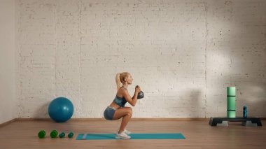 Evde online olarak kişisel spor dersleri. Spor giyim bölümünde egzersiz yapan sarışın bir kadın. Sağlık hizmetleri yaratıcı reklam konsepti. Kadın fitness koçu odanın yan tarafında kettlebell ile çömelmiş duruyor..