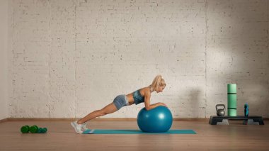 Evde online olarak kişisel spor dersleri. Spor giyim bölümünde egzersiz yapan sarışın bir kadın. Sağlık hizmetleri yaratıcı reklam konsepti. Odadaki kadın fitness koçu topun üzerine tahta çakıyor..