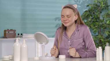 Yüzüne krem sürmüş bir kadın, gözleri kapalı oturur ve yumruklarını sıkarak sonucu bekler. Saç bandı ve ipek pijama giyen genç bir kadın sabahları...