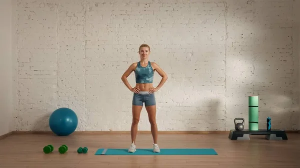 Personal sport classes at home online. Blonde female in sportswear doing exercises. Healthcare creative advertisement concept. Woman fitness coach in the room smiling looking at the camera.
