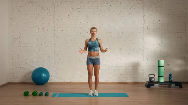 Personal sport classes at home online. Blonde female in sportswear doing exercises. Healthcare creative advertisement concept. Woman fitness coach in the room looking at the camera and talking.