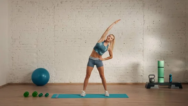 Personal sport classes at home online. Blonde female in sportswear doing exercises. Healthcare creative advertisement concept. Woman fitness coach in the room doing side body tilt stretching.