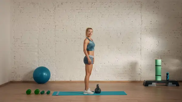 Personal sport classes at home online. Blonde female in sportswear doing exercises. Healthcare creative advertisement concept. Woman fitness coach in the room looking smiling, kettlebell on the floor.