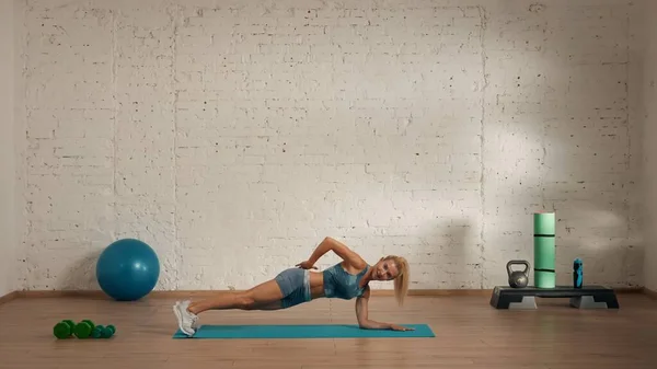 Personal sport classes at home online. Blonde female in sportswear doing exercises. Healthcare creative advertisement concept. Woman fitness coach in the room showing static plank technique.