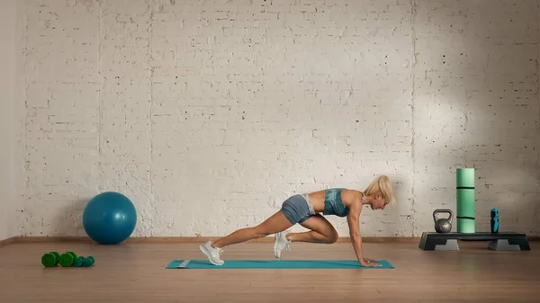 Personal sport classes at home online. Blonde female in sportswear doing exercises. Healthcare creative advertisement concept. Woman fitness coach in the room doing running in plank.