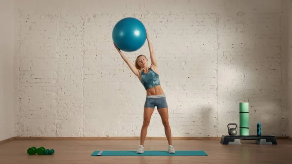Personal sport classes at home online. Blonde female in sportswear doing exercises. Healthcare creative advertisement concept. Woman fitness coach in the room doing side tilts with fit ball.