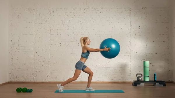 Personal sport classes at home online. Blonde female in sportswear doing exercises. Healthcare creative advertisement concept. Woman fitness coach in the room doing lunges with fit ball.