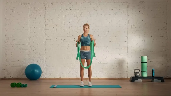 Personal sport classes at home online. Blonde female in sportswear doing exercises. Healthcare creative advertisement concept. Woman fitness coach in the room talking about rubber band.
