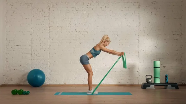 Personal sport classes at home online. Blonde female in sportswear doing exercises. Healthcare creative advertisement concept. Woman fitness coach in the room doing rubber band half bend lifts.