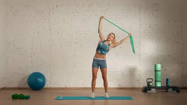 Personal sport classes at home online. Blonde female in sportswear doing exercises. Healthcare creative advertisement concept. Woman fitness coach in the room doing rubber band arm stretch.
