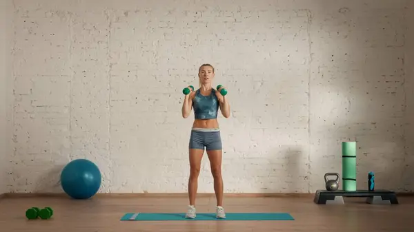 Personal sport classes at home online. Blonde female in sportswear doing exercises. Healthcare creative advertisement concept. Woman fitness coach in the room holding dumbbells on shoulders.