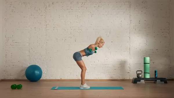 Personal sport classes at home online. Blonde female in sportswear doing exercises. Healthcare creative advertisement concept. Woman fitness coach in the room doing dumbbells half bend lift.