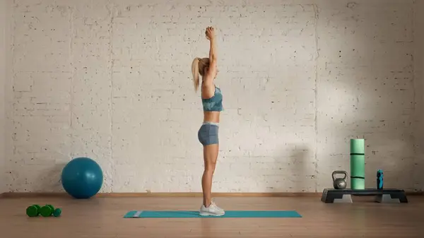 Personal sport classes at home online. Blonde female in sportswear doing exercises. Healthcare creative advertisement concept. Woman fitness coach in the room holding dumbbell in raised hands.