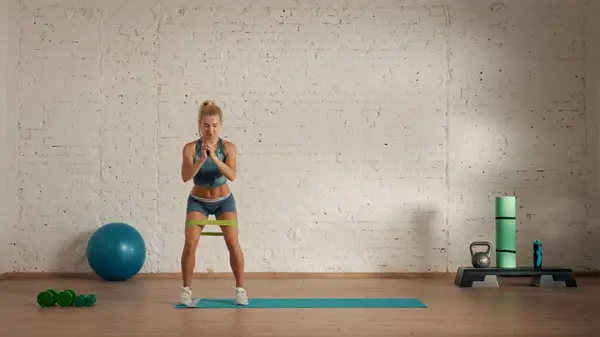 Personal sport classes at home online. Blonde female in sportswear doing exercises. Healthcare creative advertisement concept. Woman fitness coach in the room doing rubber band side steps.