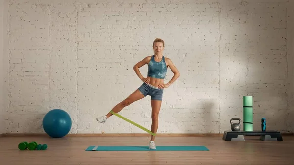 Personal sport classes at home online. Blonde female in sportswear doing exercises. Healthcare creative advertisement concept. Woman fitness coach in the room doing rubber band left leg rise.