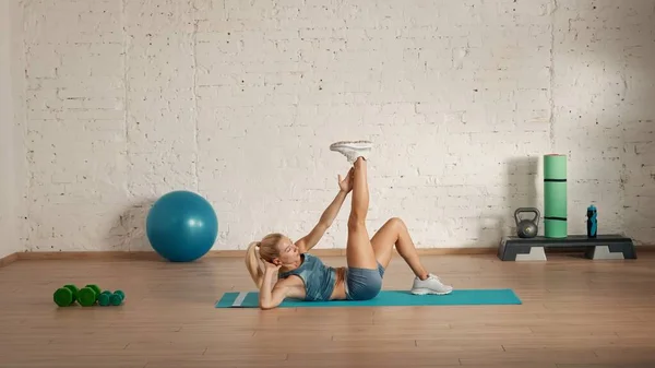 Personal sport classes at home online. Blonde female in sportswear doing exercises. Healthcare creative advertisement concept. Woman fitness coach in the room doing abs exercise with leg up.