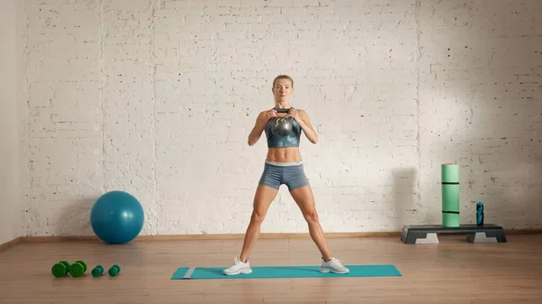 Personal sport classes at home online. Blonde female in sportswear doing exercises. Healthcare creative advertisement concept. Woman fitness coach in the room standing in wide position with kettlebell