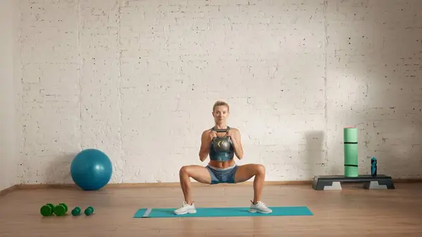 Personal sport classes at home online. Blonde female in sportswear doing exercises. Healthcare creative advertisement concept. Woman fitness coach in the room doing squats in wide with kettlebell.