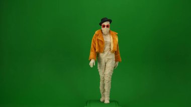 Turuncu ceketli, şapkalı ve güneş gözlüklü göz kamaştırıcı mumya kameraya doğru zarifçe yürüyor. Yeşil ekran krom anahtarı tam boy fotoğraf, modelleme, reklam için çalışma alanı. Tam uzunluk.