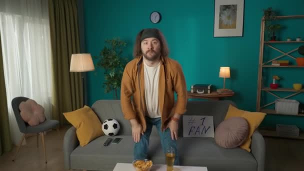 画面显示一个中年男子坐在沙发上吃薯片 在电视上观看足球比赛的演示者 他站起身来 用手捂住心脏 然后坐下来吃薯片 — 图库视频影像