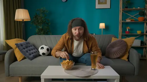 A middle aged man is sitting on a couch in an apartment. Next to him is a beer and chips, he reaches for chips and looks at the camera with a surprised look. Demonstrates Watching Football.