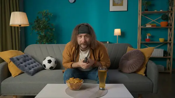 A middle aged man is sitting on a couch in an apartment. Next to him is a beer and chips, he reaches for chips and looks at the camera with a surprised look. Demonstrates watching football.