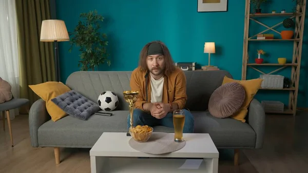 In the shot, a middle aged man sits on a sofa in the living room. Next to him is a ball, on the table is a gold cup, beer and chips. He gazes at the cross arms camera. He is focused, attentive.