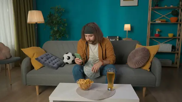 ショットでは 中年男性がソファーに座っています 彼は見ているドルと電話のファンを持っている ボールとビールとチップが隣に横たわっている サッカーファンを模倣する — ストック写真
