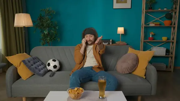 フレームは アパートのソファーに座っている中年の男性を示しています 彼は電話で誰かと話し テレビのようにカメラを覗いている 彼はサッカーの試合について話している — ストック写真