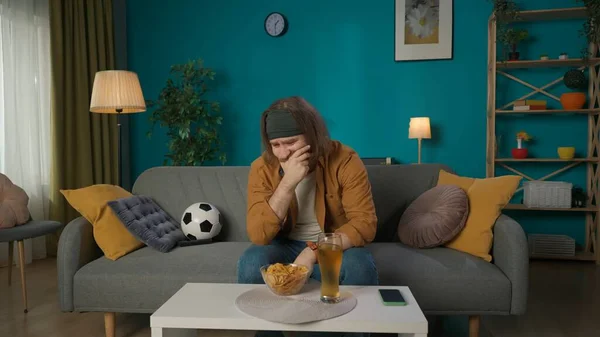 在照片中 一个中年男人正坐在房间的沙发上 足球旁边是啤酒和薯片 他面无表情地望着别处 打了一个足球迷 看起来很不高兴 很伤心 — 图库照片