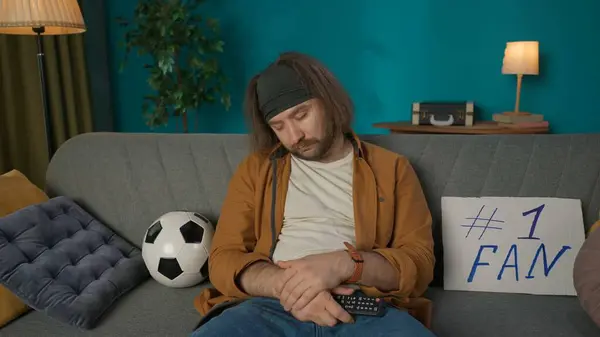 中年男性が部屋のソファーに座っています 彼の隣にはサッカーボールとタブレットがある 彼はテレビのリモコンを持って眠ってしまった サッカーを見ながら眠りに落ちるファンを肖像画 — ストック写真