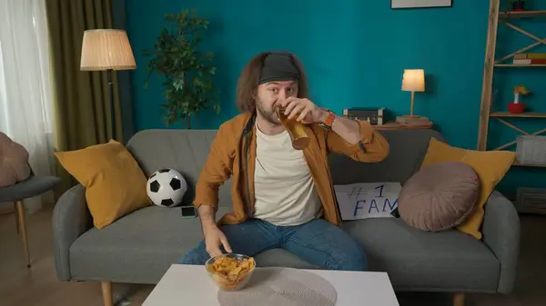 中年男性がソファーに座っています 彼の隣にはファンタブレットがあり テーブルにはビール チップ 電話があります カメラを見てビールを飲む 試合を見ているサッカーファンを肖像画 — ストック写真