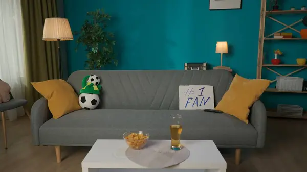 In the frame there is an empty living room with a cozy atmosphere. In the middle of the room there is a gray sofa on which there is a football paraphernalia, a ball, a fan hat and a TV remote control.