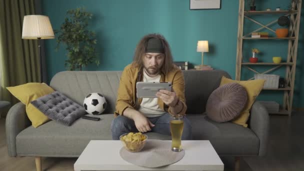 프레임은 소파에 앉아있는 남자를 보여줍니다 태블릿을 경기를 긴장과 먹고있다 그리고 — 비디오