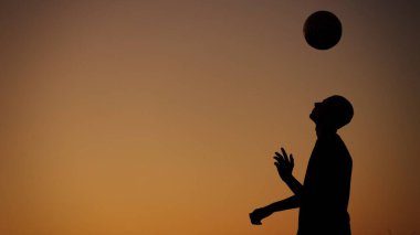 Gün batımında dışarıda topla oynayan genç bir adamın siluetini gösteren orta boy bir fotoğraf. O, topu havaya fırlatıyor. Spor, futbol ve diğer sporlar, gençlik.