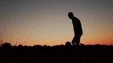 Gün batımında tarlada topla oynayan bir gencin siluetini gösteren uzun bir video. O, topa havada tekme atıyor. Spor, futbol ve diğer sporlar, gençlik.