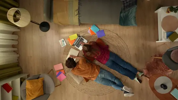 在头像中 一对年轻夫妇躺在地板上彼此相邻 在笔记本电脑上工作 做笔记 四周环绕着五彩斑斓的书籍 共度时光 — 图库照片