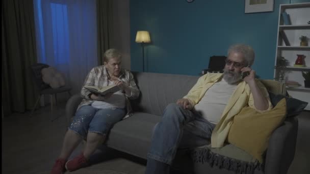 高齢のカップルがソファーに座っている その男は情熱的に電話で話している その本を読んでいる女性を予防します 彼女は気に入らない 彼女は自由ではない Hdr Bt2020 Hlg材料について — ストック動画