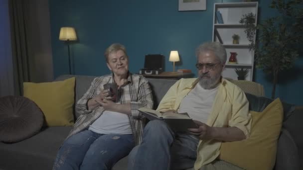 写真では 高齢のカップルは青い壁に対してアパートのソファーに座っています 男はその本を読んでいる 女性はリスニングし お茶を飲む ホーム コージーネス Hdr Bt2020 Hlgについて — ストック動画