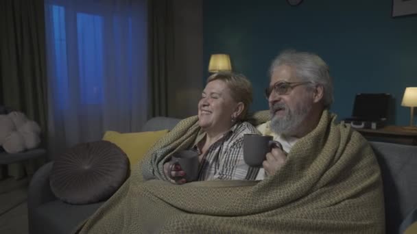 写真では 高齢のカップルが毛布の下の部屋のソファーに座っています 彼らは熱い紅茶やコーヒーを飲んでいる テレビで映画や放送を見る Hdr Bt2020 Hlg材料について — ストック動画