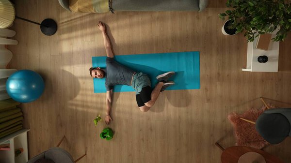 Вид сверху на молодого человека, делающего утреннюю зарядку, растянувшегося перед окном. Дом, комната, занятия в помещении. Здоровый и активный образ жизни, утренняя рутина, достижение целей.