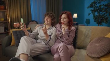 Aile ve ilişkiler reklam konsepti. Birlikte zaman geçiren genç bir çiftin portresi. Kanepede pijamalı bir adam ve kadın lolipop yiyor, akıllı telefondan selfie çekiyor..