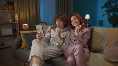 Aile ve ilişkiler reklam konsepti. Birlikte zaman geçiren genç bir çiftin portresi. Kanepede pijamalı bir adam ve kadın lolipop yiyor, tabletten video ile konuşuyor..