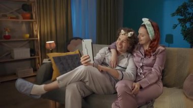 Aile ve ilişkiler reklam konsepti. Birlikte zaman geçiren genç bir çiftin portresi. Pijamalı bir adam ve kadın, saç bantlarıyla kanepede oturmuş tabletteki video konuşmalarıyla konuşuyorlar..